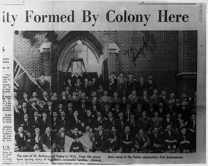 Clip from newspaper article, Hamilton Spectator, featuring photograph of parishioners of St. Anthony of Padua, Hamilton, ON, 1912. Photograph shows five rows of men standing outside a building exterior, presumably the church. A large union jack flag appears in the left background. The caption reads, "The men of St. Anthony's of Padua in 1912. From this group have sprung many of Hamilton's successful families. Among them some of the Italian community's first businessmen."