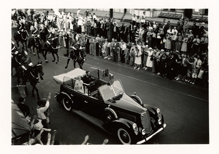 Black and white photograph of the royal motorcade with King George VI and Queen Elizabeth during the Royal Visit through Niagara Falls, ON on June 7, 1939. The vehicle is a 1939 Lincoln convertible sedan. The King and Queen are being accompanied by the Royal Canadian Dragoons (RCD), the most senior cavalry regiment in Canada. Crowds of onlookers line both sides of the street. The 1939 Royal Visit was the first visit of the reigning monarch to Canada. Large crowds greeted the royal couple throughout the cross-country tour. The tour began in May of 1939 in Quebec City. From here the King and Queen travelled west by rail, visiting most of the major cities and finally arriving in Vancouver. Then they travelled through the United States, along with Prime Minister Mackenzie King. The tour ended with a visit to the Maritimes and Newfoundland, departing from Halifax. On June 7, 1939 the Royals passed through Southern Ontario on their way to Washington, DC for a meeting with President Roosevelt. It would be the first state visit to the United States by a reigning British monarch.